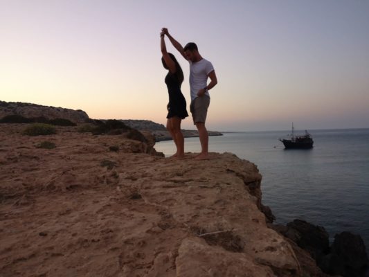 junges Pärchen tanzt im Sonnenuntergang auf einer Klippe. rechts davon liegt weiter unten das Meer